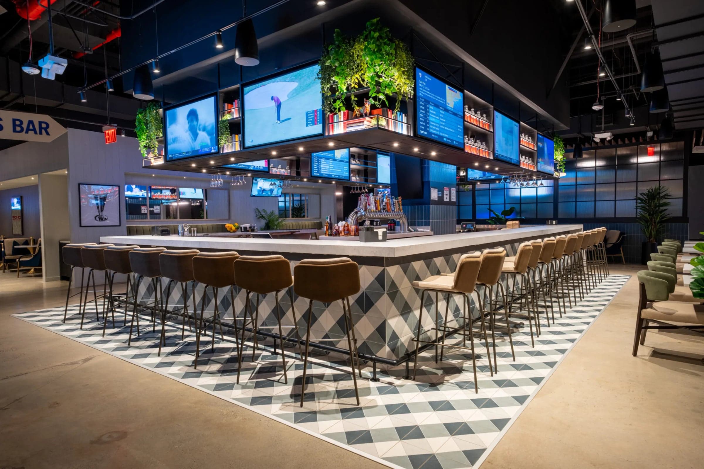 GolfZon Social Brooklyn — Main bar under a wall of screens: checkerboard rug, brass-and-cane stools, planting overhe