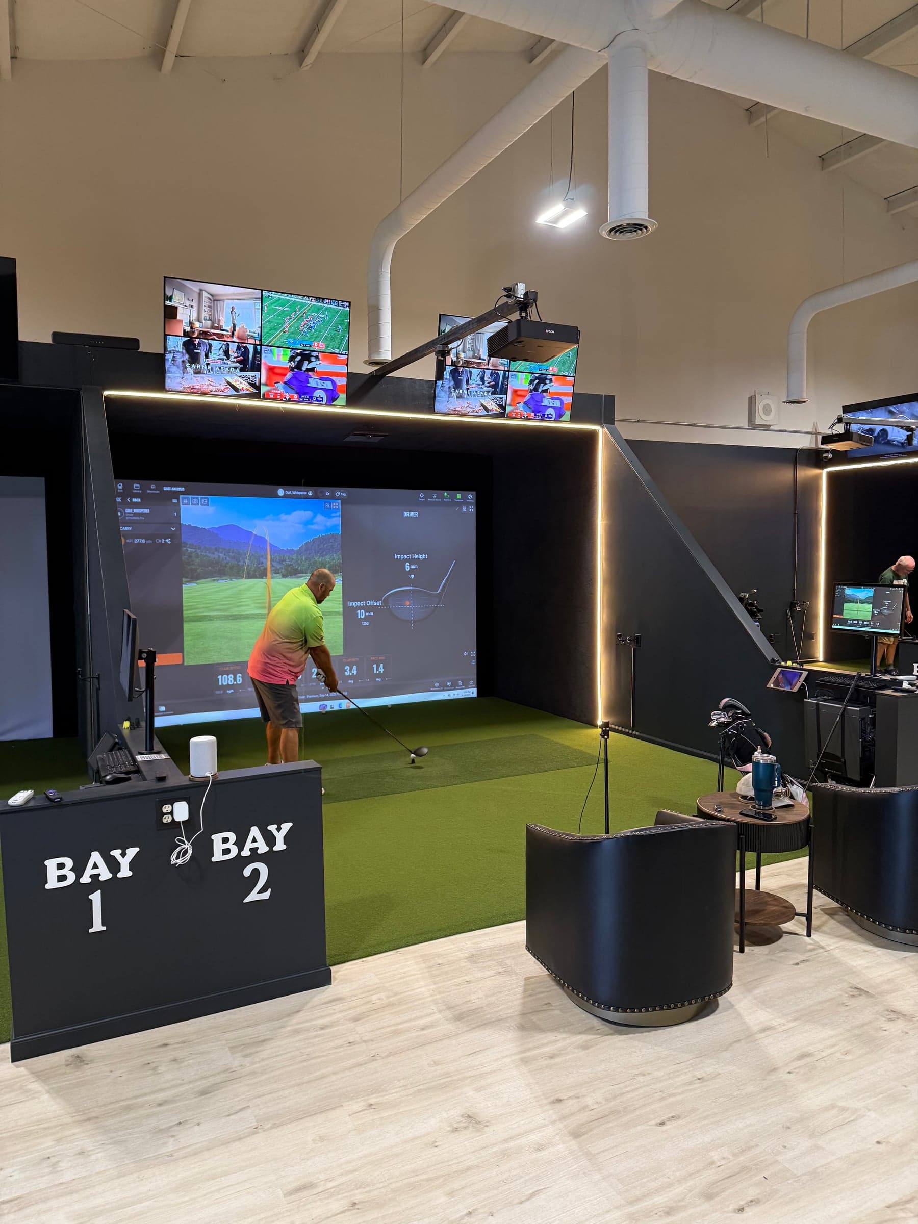 Ohio Golf Lounge — BAY 1 / BAY 2 signage on a black podium, LED-strip-lit black bays, pale timber floor, golf